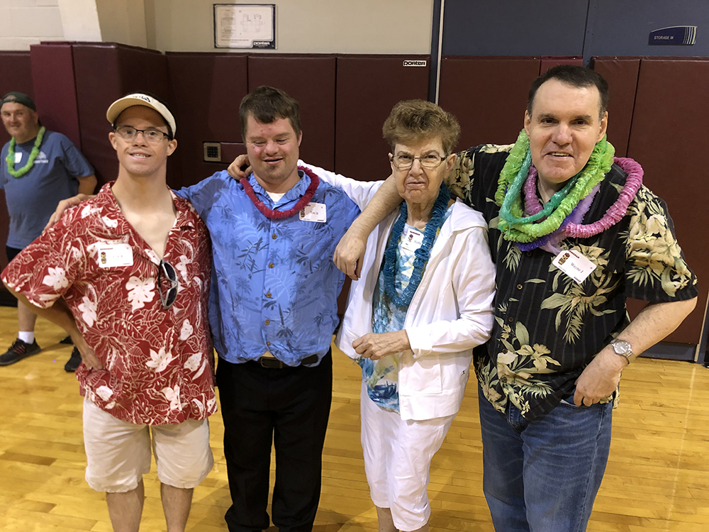 Four people, all wearing Hawaiian themed shirts, stand and happily pose together for the picture in what looks to be a gym.