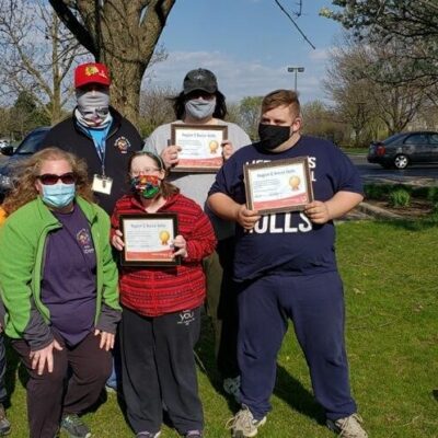 6 people, all wearing medical face masks, stand and pose together outside. 3 of them are holding up awards and 1 award is lying on the grass.