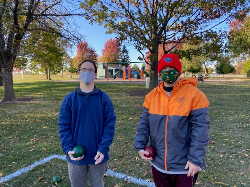 2 men pose for the photo together while holding green and red balls in a park. There is a playground in the background. The sky is clear and bright.