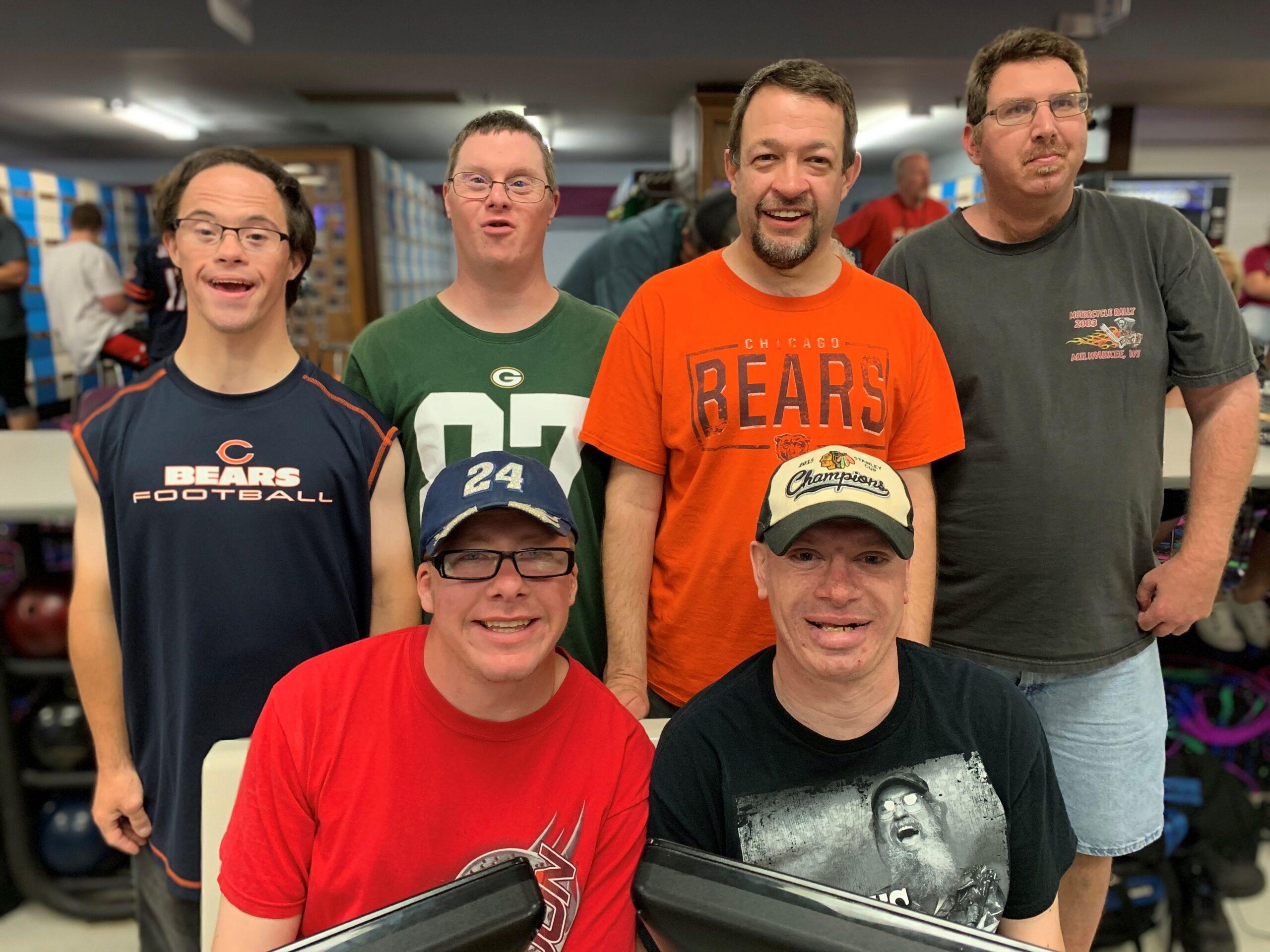 Six men smile and pose together at a bowling alley. Two of them are sitting down and the rest are standing behind them. There are bowling balls and several other people in the background.