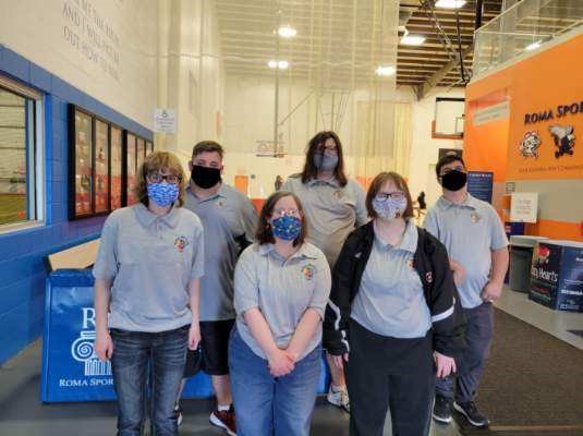 6 people, all wearing medical face masks and matching grey shirts, stand together for the photo. They are at a sports complex to take part in an activity day.