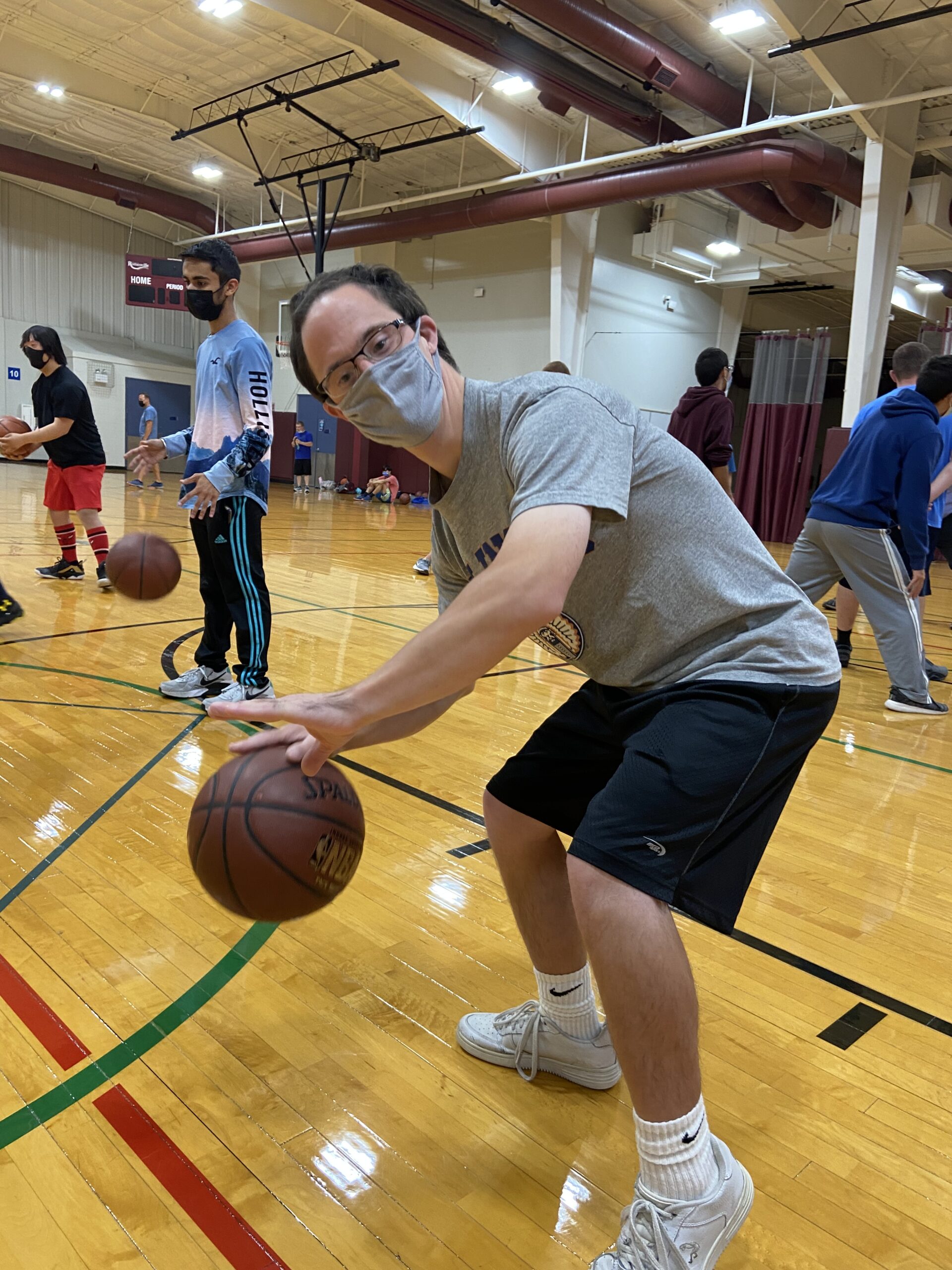 A man wearing a medical face mask dribbles a basketball on the court as he poses for the picture. There are several people in the background also playing basketball.