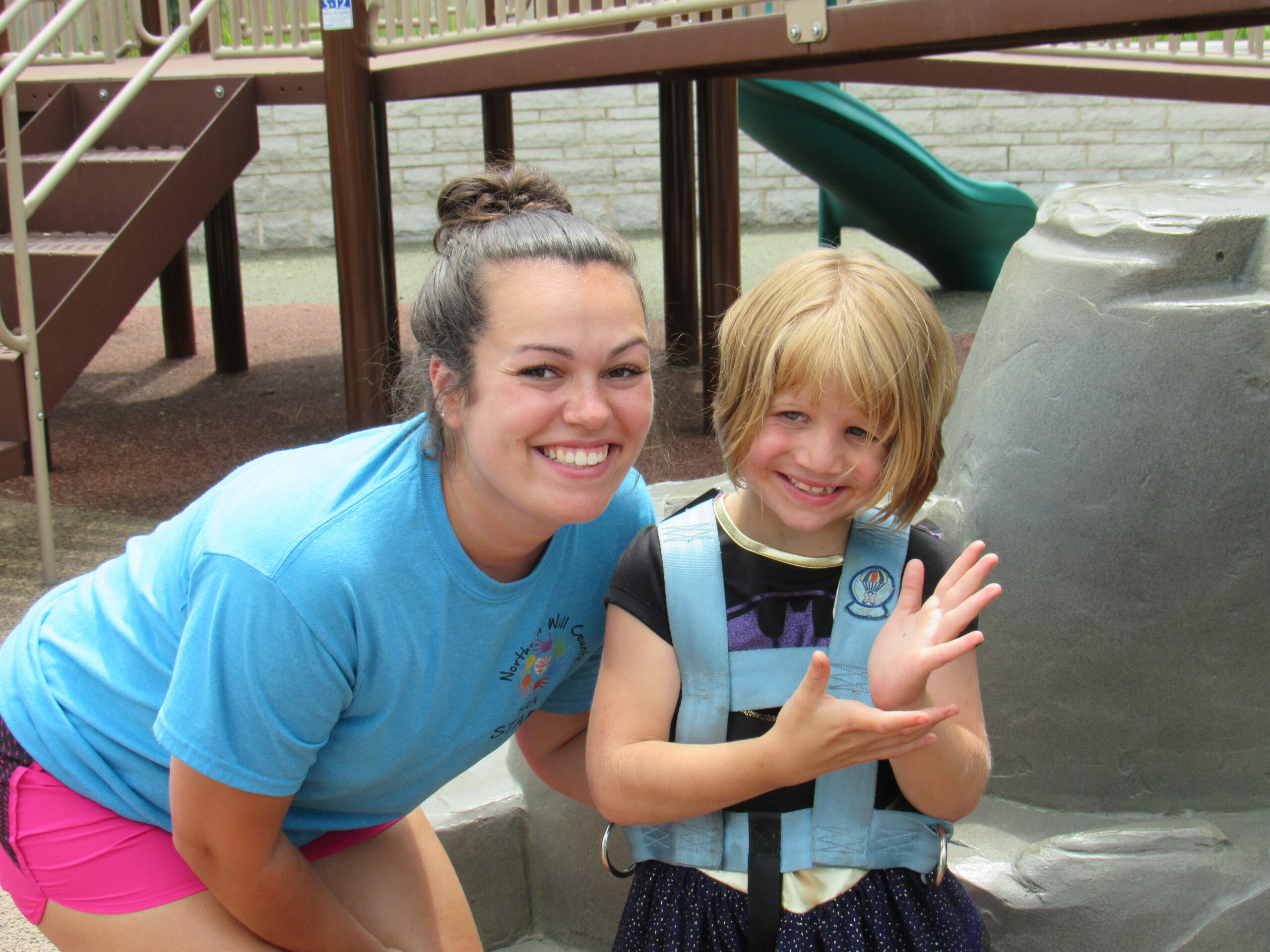 A woman and a girl stand together and smile while posing for the picture. They are at a playground and there are plastic climbable rocks and a jungle gym directly behind them.