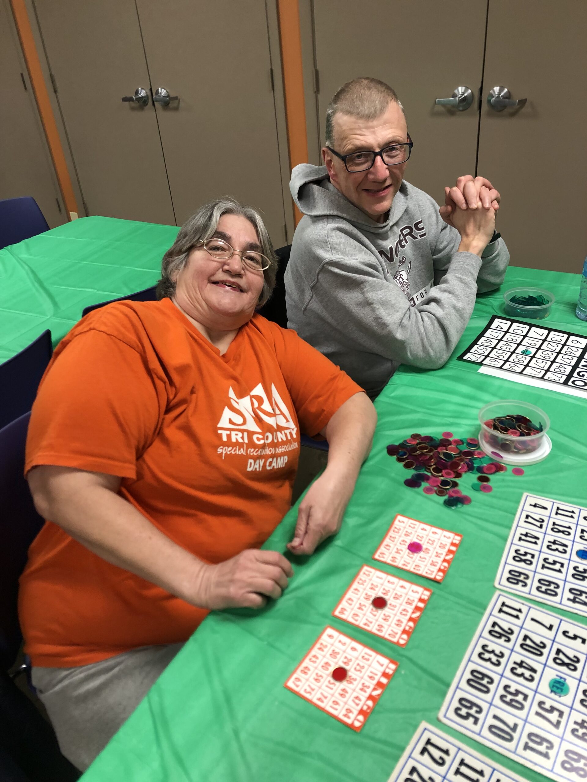 2 adults smile and pose for the picture while playing Bingo. There is a few Bingo cards and a big bowl in front of them with several Bingo chips inside and on the table.