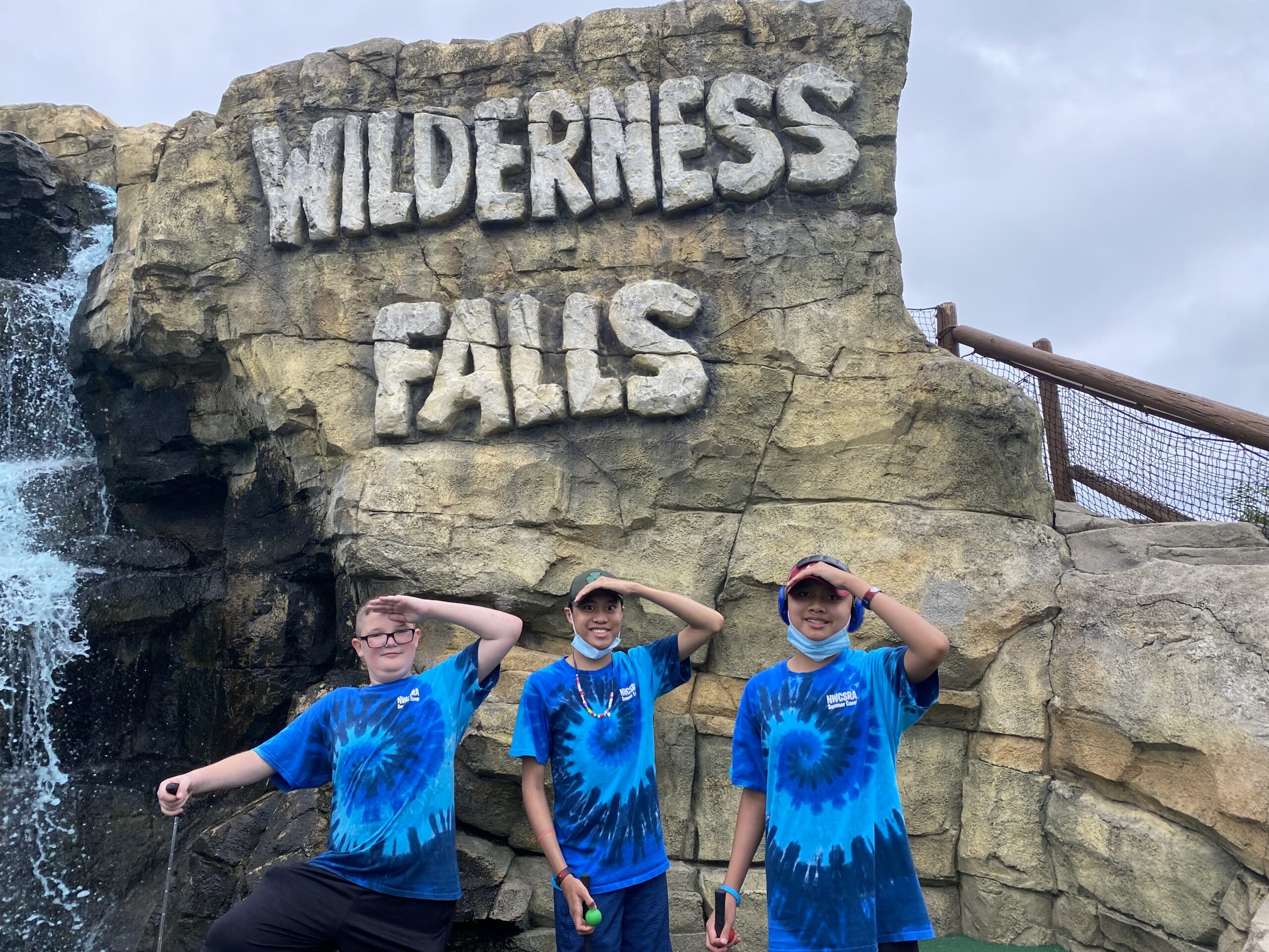 3 people, all wearing blue tie-dye shirts that say ‘NWCSRA Summer Camp’, pose in front of a rock wall that says ‘Wilderness Falls’. To the left of where they’re standing is a small waterfall.